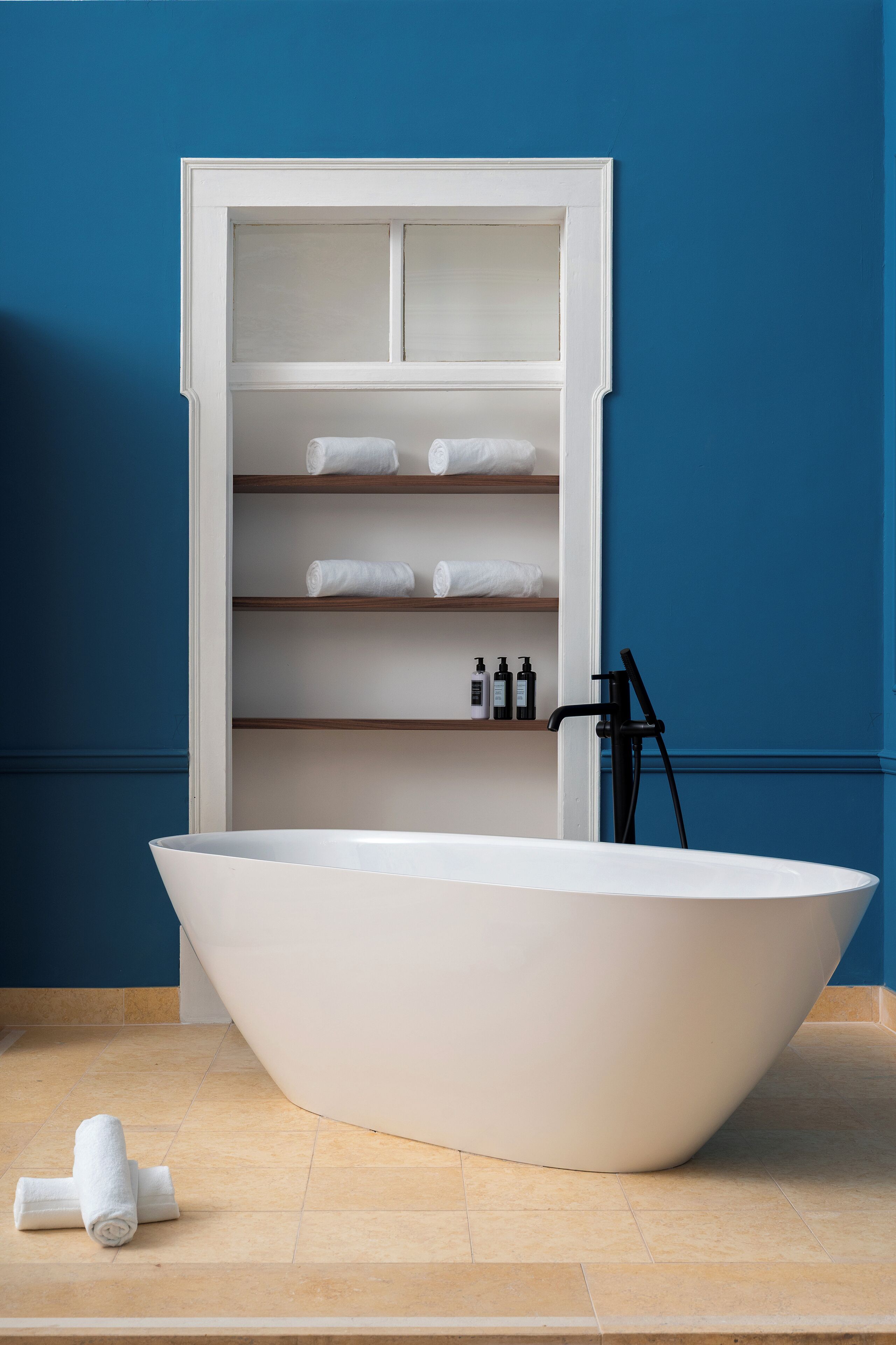A freestanding bathtub in a bathroom with a built-in shelf unit above it, holding towels and toiletries.