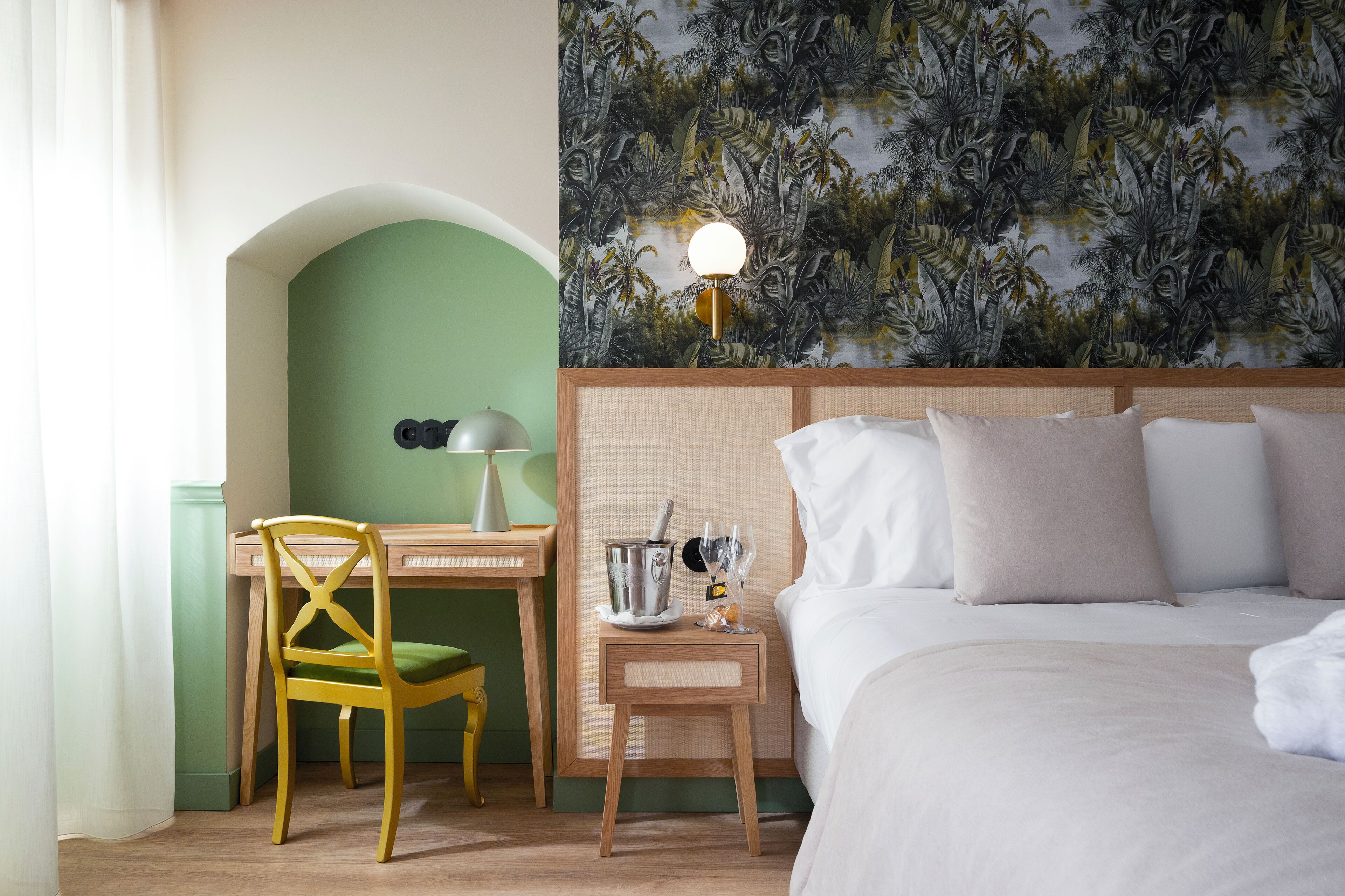 A hotel room with a bed, a desk with a chair, a nightstand with a lamp, and a wall with a tropical wallpaper.