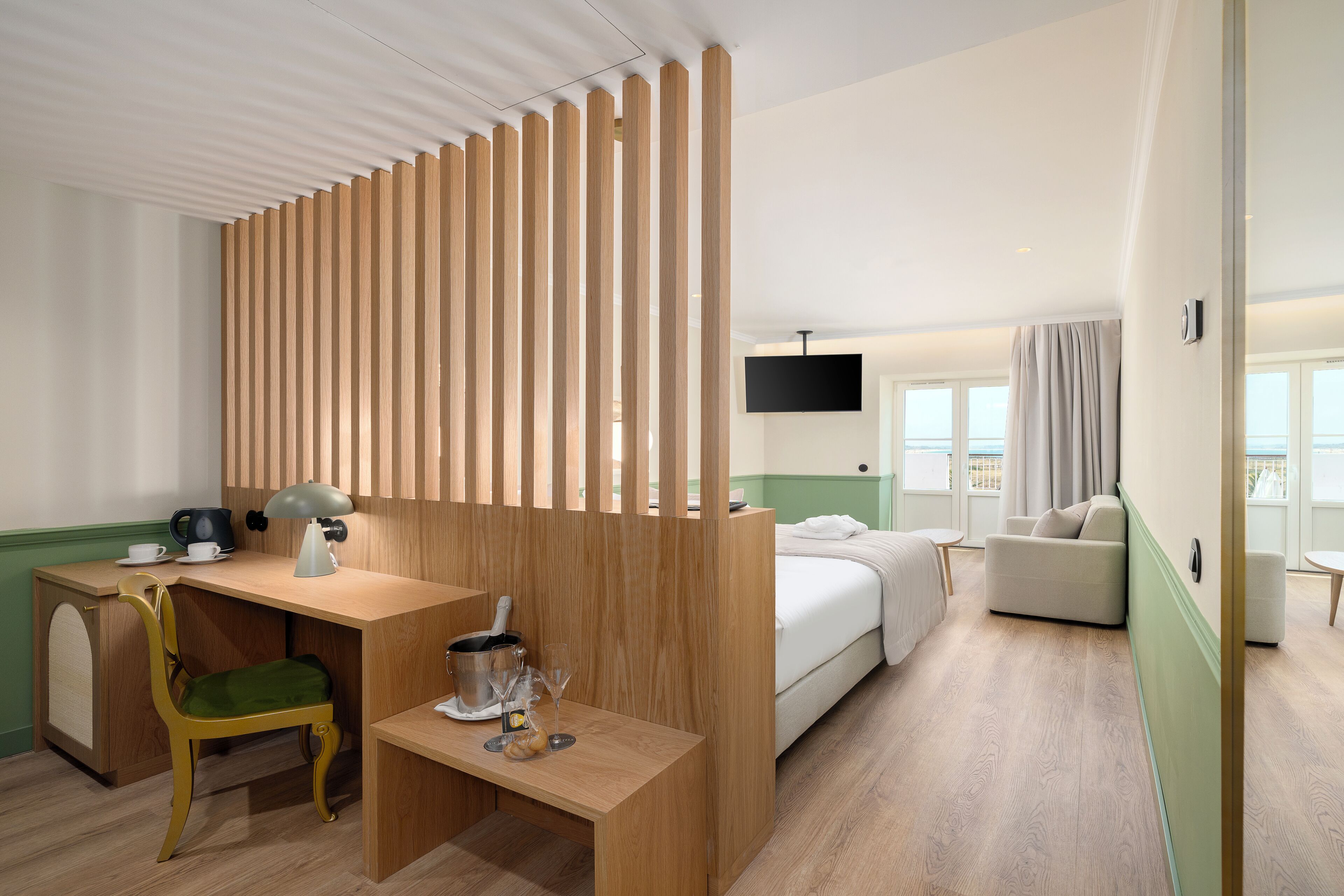 A hotel room with a wooden desk, a green chair, a bed, a TV mounted on the wall, and a view of the outside through large windows.