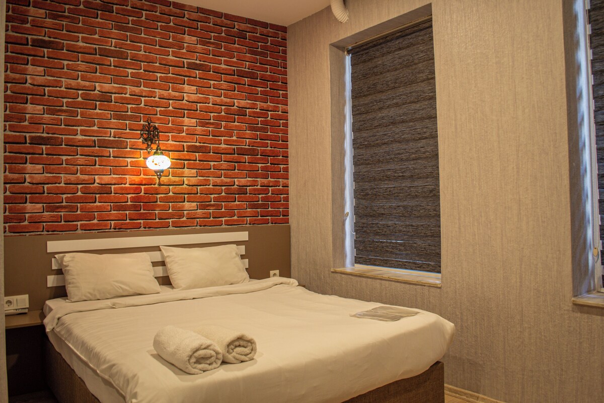 Una camera d'albergo con una parete in mattoni, un'ampia finestra con persiane, un letto con lenzuola bianche e un lampadario fissato al muro.