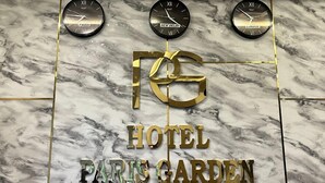 Une enseigne d’hôtel avec trois horloges indiquant des heures différentes, et le nom de l’hôtel, « HOTEL PARIS GARDEN », affiché en grand.