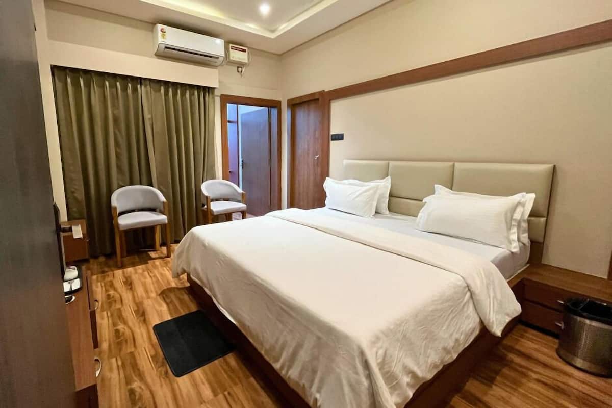 A hotel room with a large bed, two chairs, a desk, an air conditioning unit, and a trash can.