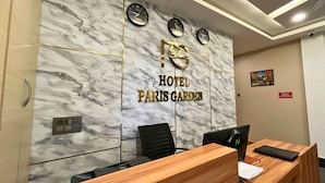 Un comptoir d’accueil d’hôtel avec un mur en marbre, des horloges et une enseigne indiquant « HÔTEL PARIS GARDEN ».