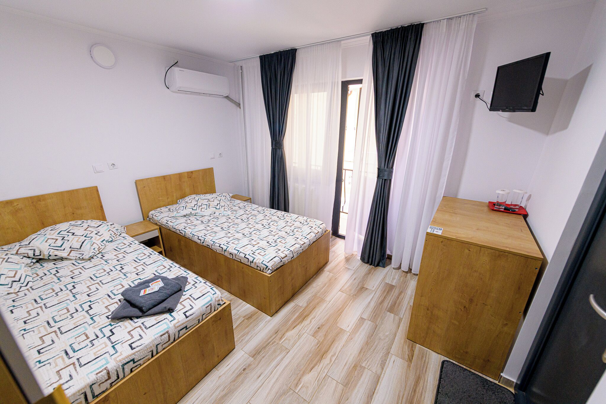 Una habitación de hotel compacta con dos camas individuales, un escritorio de madera, un televisor montado en la pared y una ventana con cortinas.