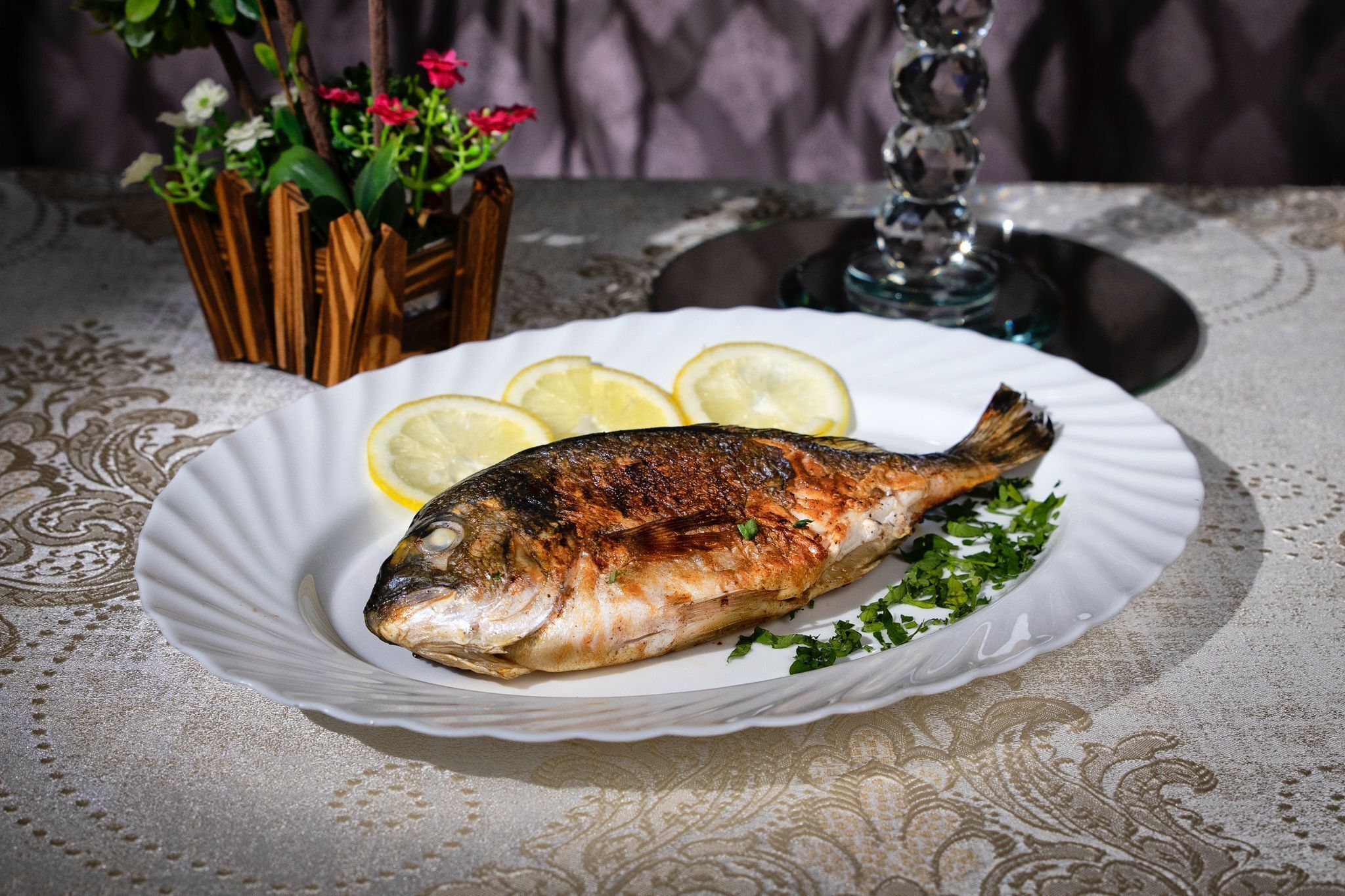 Un pescado a la parrilla en un plato con rodajas de limón y perejil, sobre una mesa con mantel de encaje.