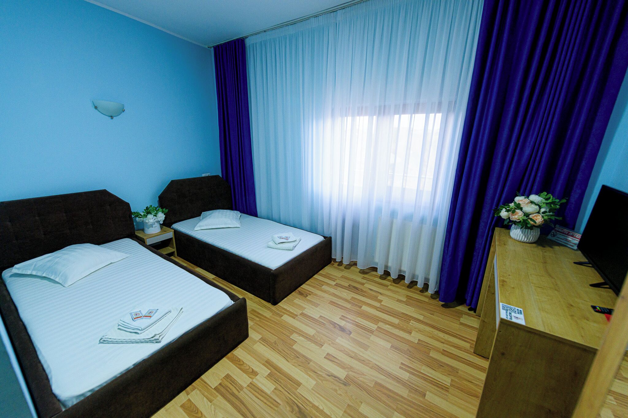 A hotel room with two single beds, a wooden desk with a TV, and a window with purple curtains.