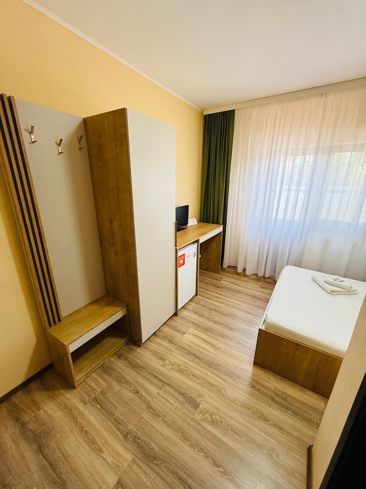 A compact hotel room with a bed, a desk with a computer, a wardrobe, and a small seating area.