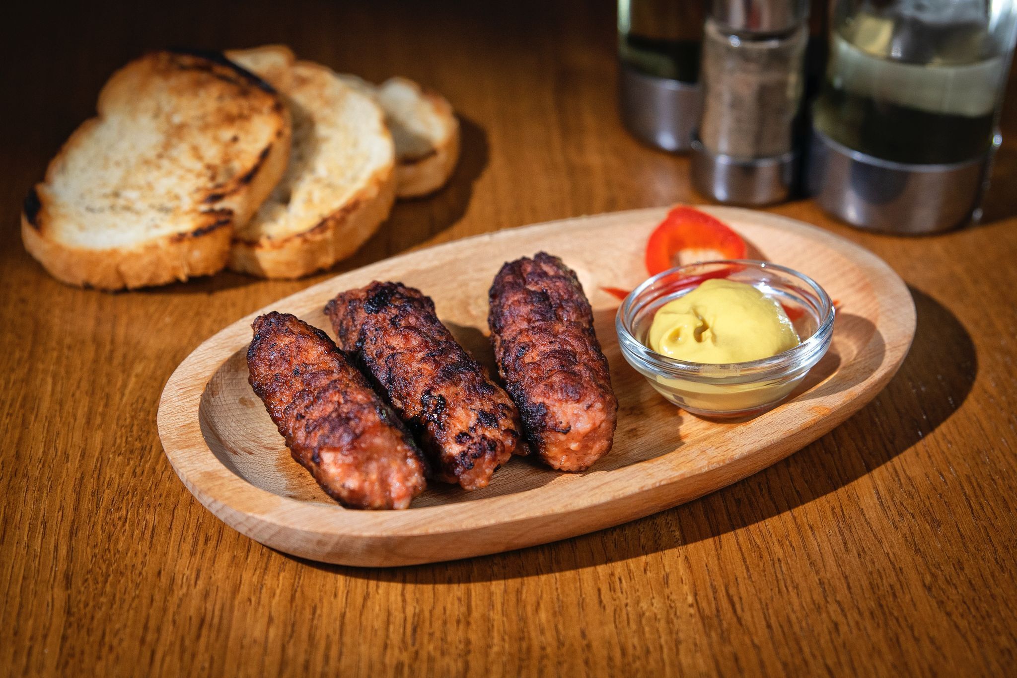 A wooden plate with grilled sausages, a small bowl of mustard, and toasted bread on a wooden table.