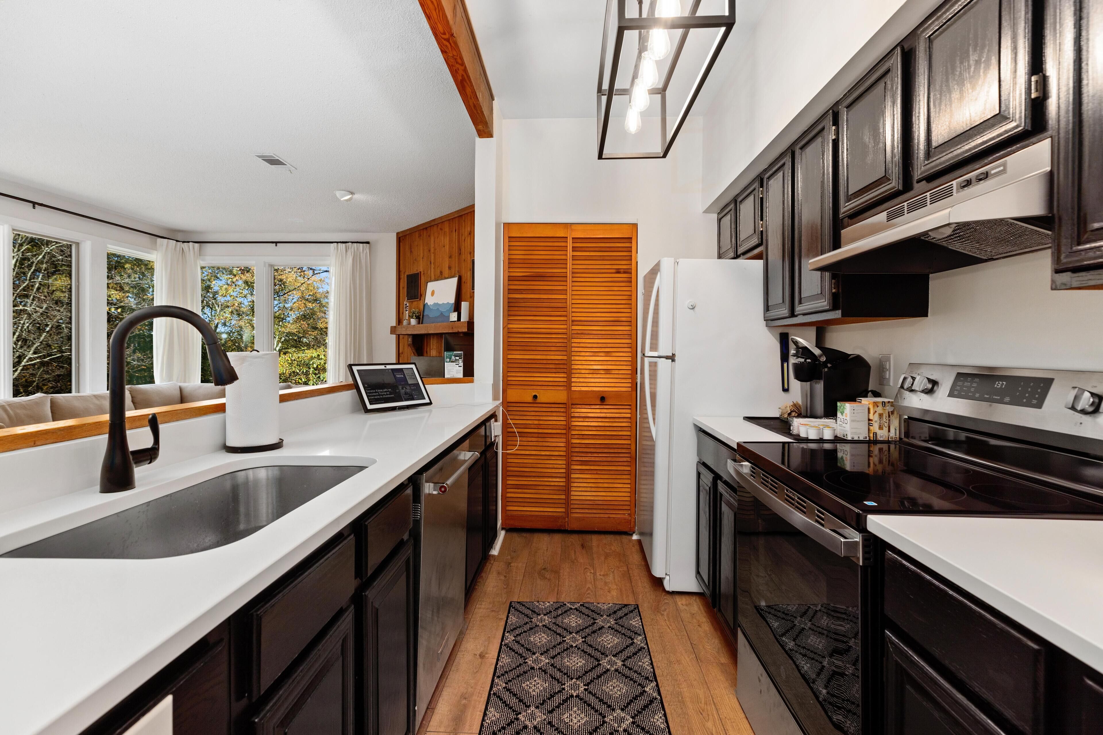 A modern kitchen with dark wooden cabinets, white countertops, a built-in oven, and a large window providing a view of the outdoors.