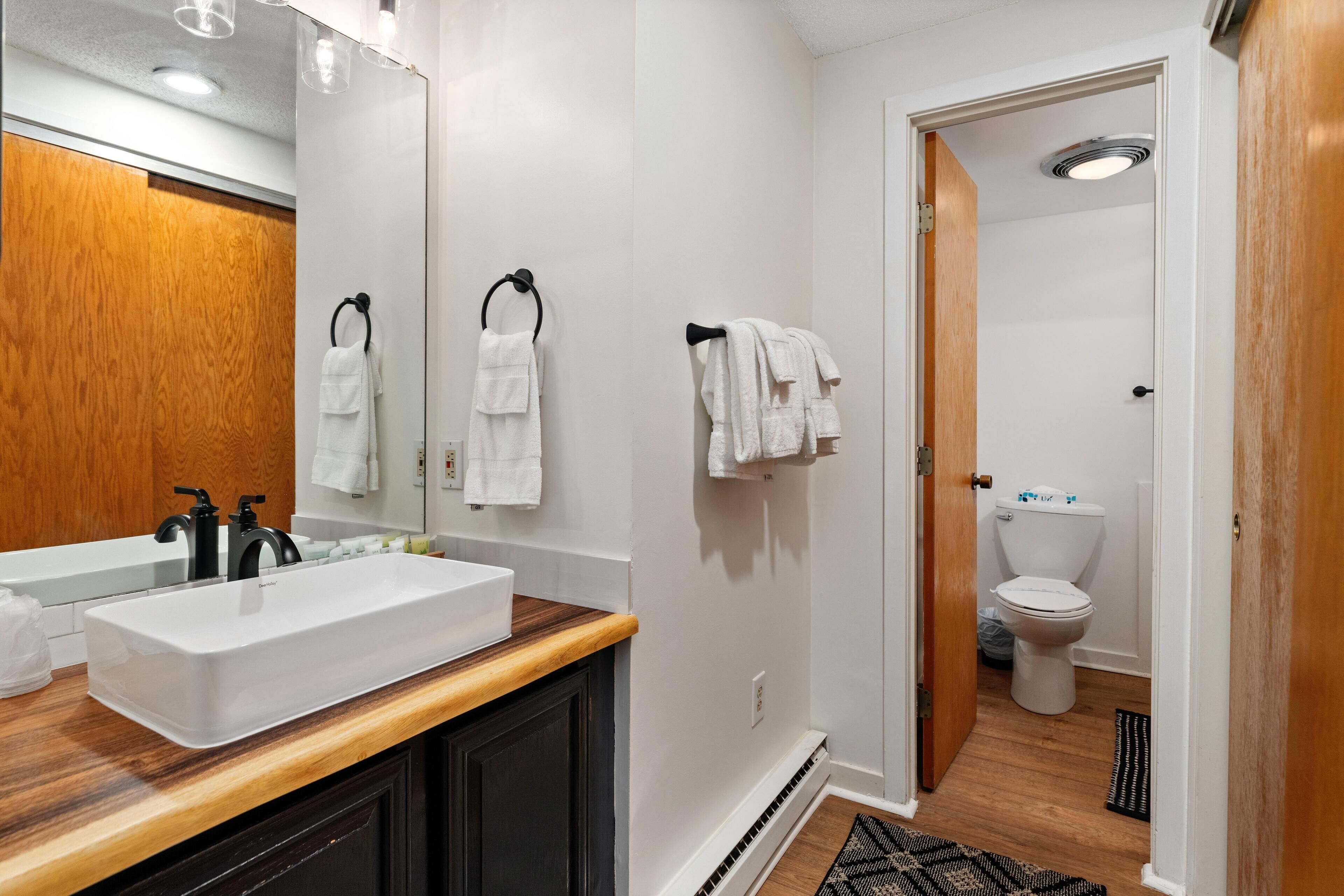 A modern bathroom with a large white sink, black faucets, and a wooden countertop. There are two towel rings on the wall and a door leading to the toilet.