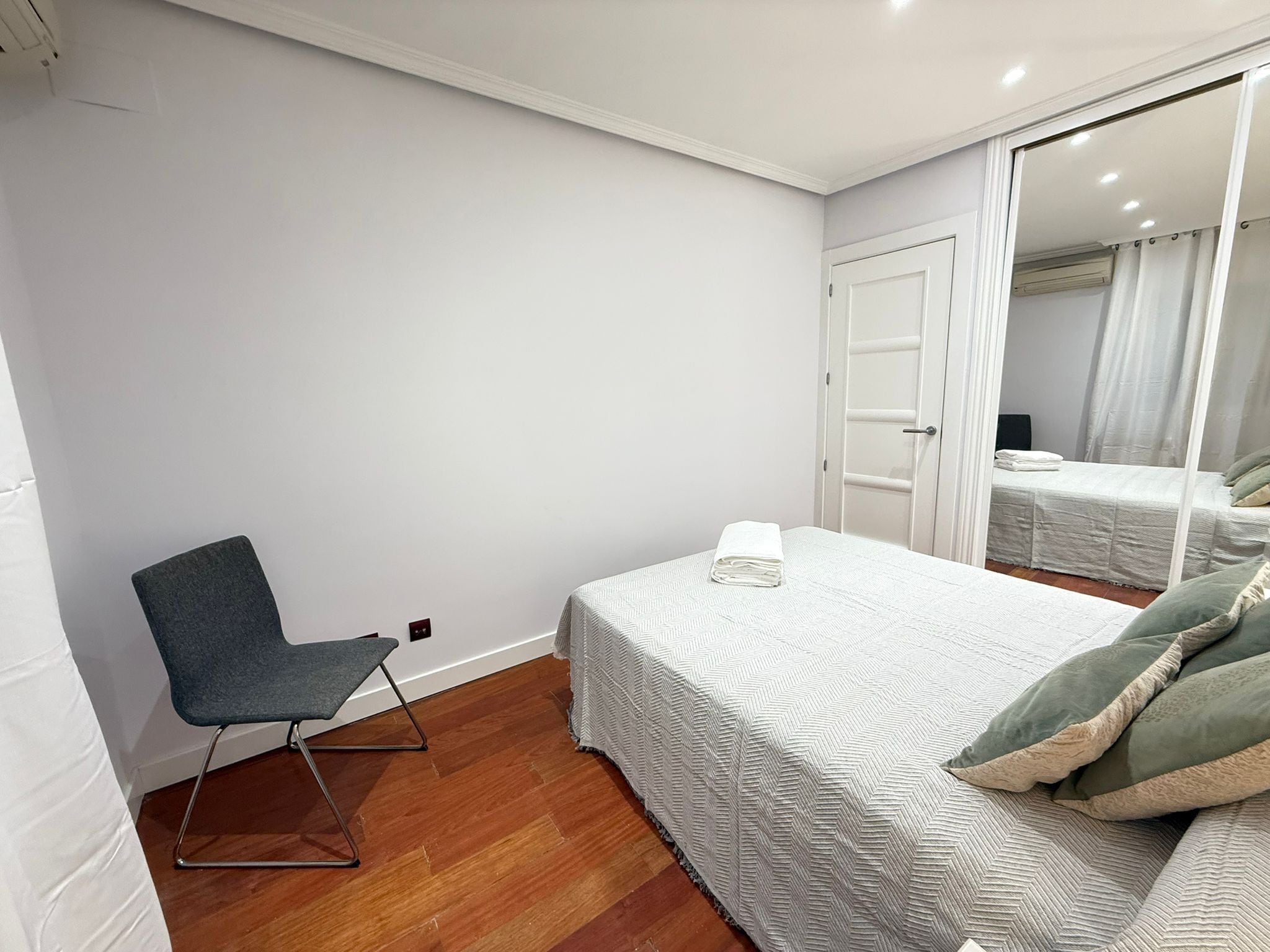 A bedroom with a bed, a chair, a bedside table, and a mirror.