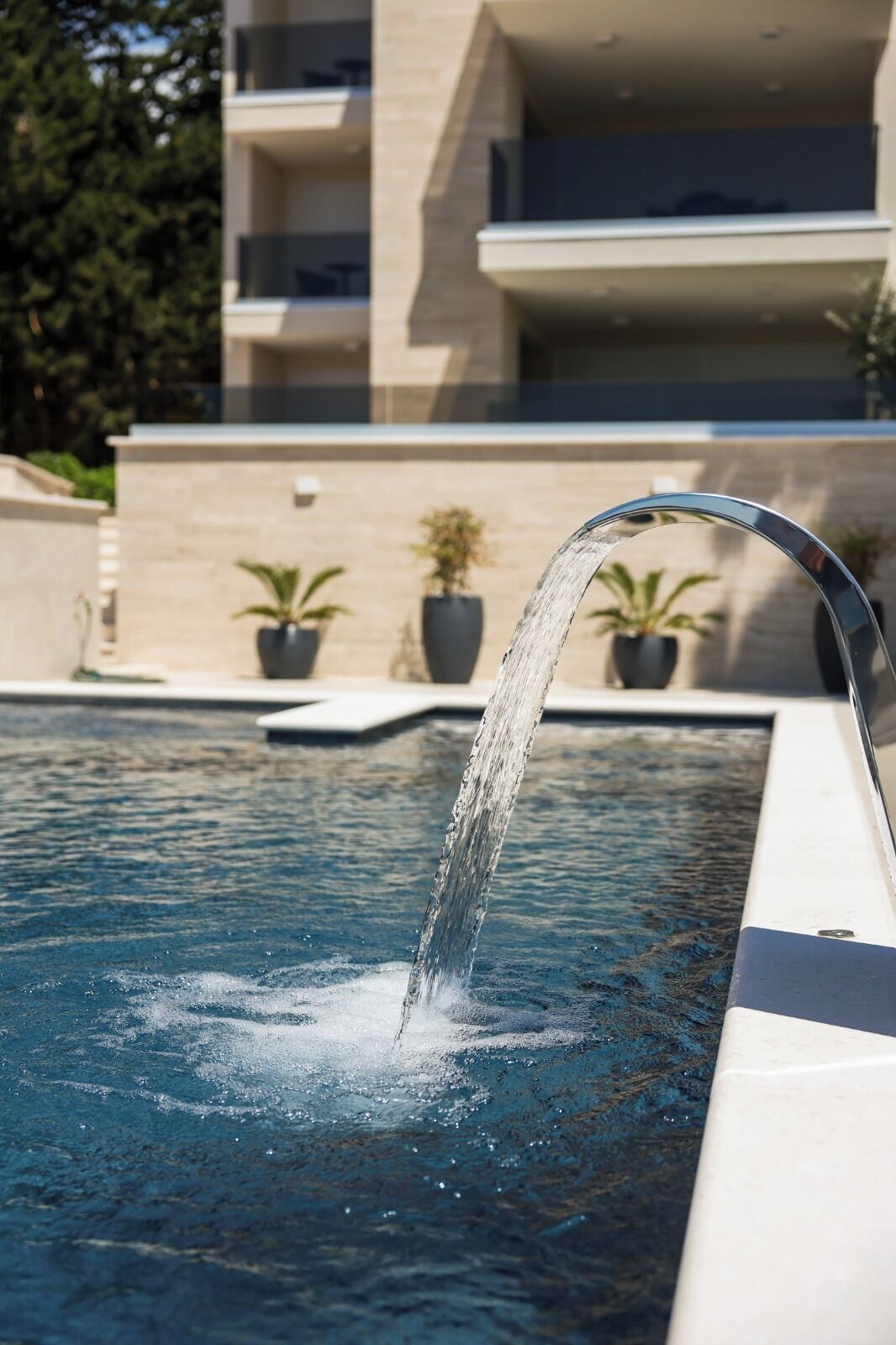 Une piscine agrémentée d’une fontaine, de plantes en pot et, en arrière-plan, d’un bâtiment moderne.