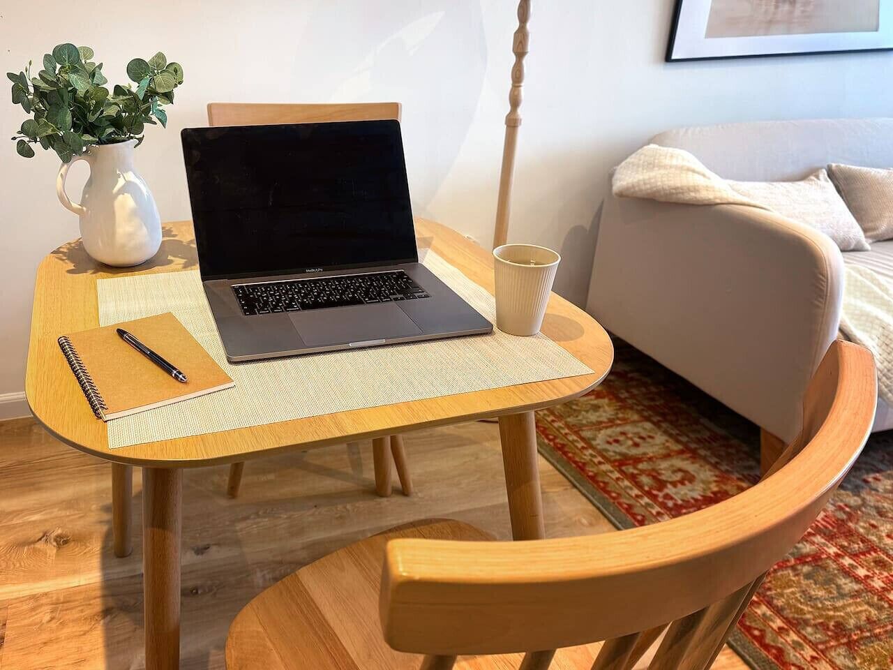 A wooden table with a laptop, a notebook, and a pen. There is a white pitcher with green plants on the table. A beige sofa and a patterned rug are in the background.
