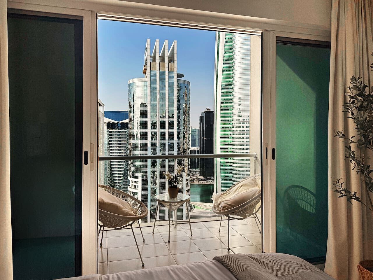 A balcony with a view of modern buildings, a small table with a plant, and two wicker chairs.