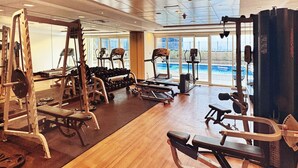 Ett gym med olika typer av träningsutrustning, ett löpband och utsikt över en pool genom stora fönster.