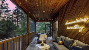 A wooden deck with a sofa and a table, a neon sign, and a view of trees. - Cedar Hill Lodge | 10 Min to Mt Snow w/Hot Tub (Wardsboro)