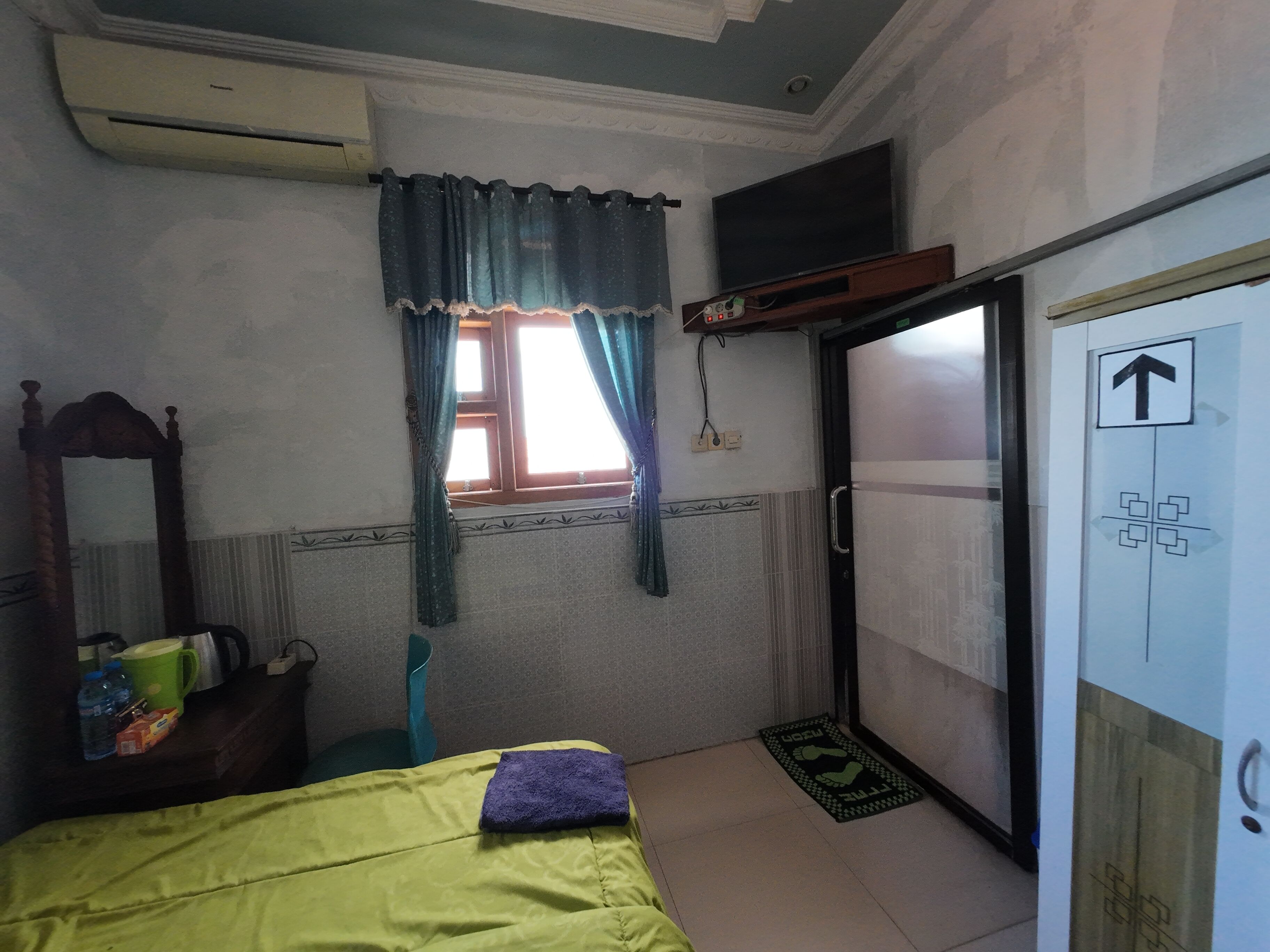 A room with a bed, a window with curtains, a mirror, a wardrobe, and a door with an arrow sign.