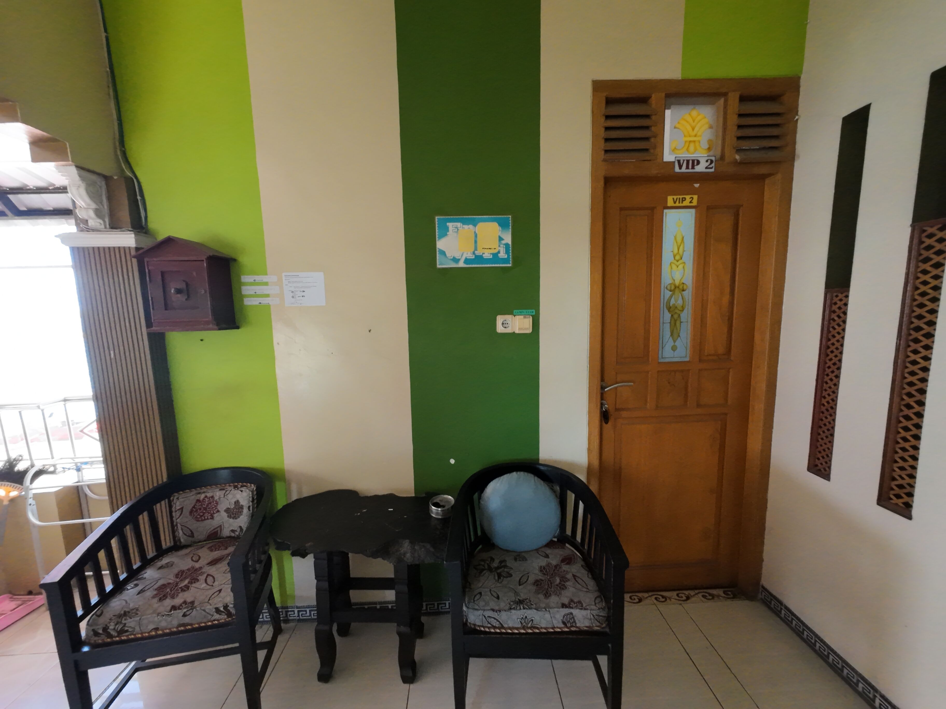 Una habitación con pared verde y blanca, una puerta de madera con un letrero que dice 'VIP 2', dos sillas con tapicería floral y una mesita con un frutero.