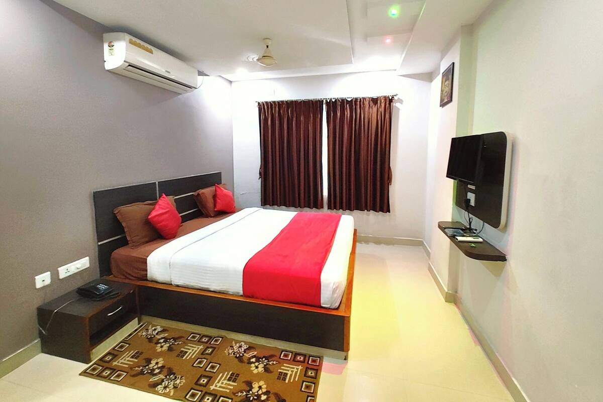 A hotel room with a bed, a TV, an air conditioner, and a window with curtains.