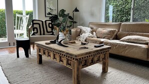 Ein Wohnzimmer mit Couchtisch aus Holz, einem beigefarbenen Sofa, einem weißen Sessel und einem großen Fenster mit Blick auf Grünflächen.