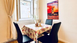 A dining area with a table set for two, a window with curtains, and a painting of a sunset on the wall. - Modern Studio Stay Near Stadthalle (Vienna)