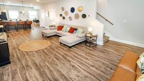 Amplia sala de estar con una cocina al fondo, un conjunto de sofás y un comedor con taburetes de bar.