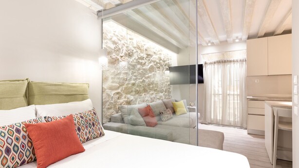A modern bedroom with a large bed, a glass partition, a stone wall, a sofa, and a kitchen area.