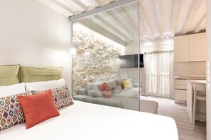 Un dormitorio moderno con una cama grande, un separador de vidrio, una pared de piedra, un sofá y una zona de cocina.