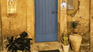 Una porta blava amb una aldreta de metall, una làmpada penjada a la paret, una planta en test i un gerro gran.