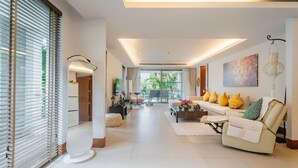A spacious living area with a sofa set, a coffee table, and a dining table. There is a large window with blinds, a ceiling light, and a wall-mounted lamp. - Elegant Beachfront 3BR Condo at Pearl of Naithon (Sa Khu)