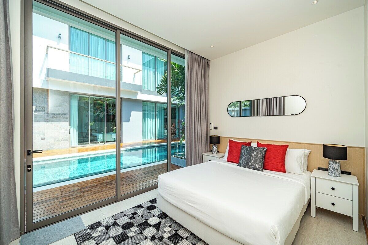 A modern hotel room with a large bed, a nightstand, a lamp, a mirror, and a view of a swimming pool through floor-to-ceiling glass doors.
