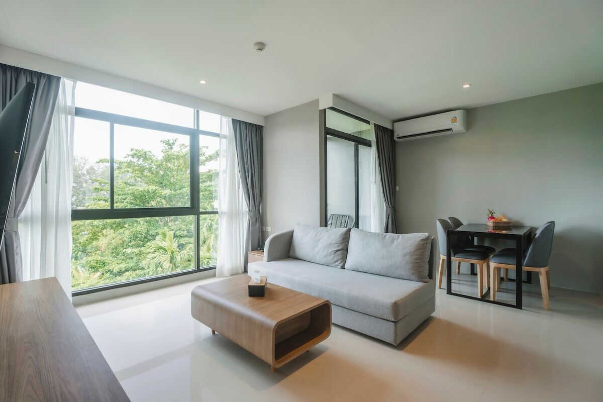 A modern living room with a grey sofa, a wooden coffee table, a dining table with chairs, and a large window with curtains.
