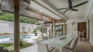 A spacious outdoor dining area with a glass table and white chairs, a ceiling fan, and a view of a swimming pool and greenery. - Luxury 3BR Villa 5 Private Pool, Gym & Jacuzzi (Choeng Thale)