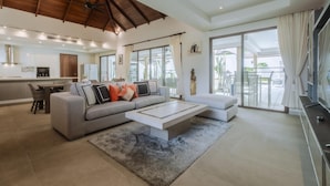 A spacious living room with a large sofa, a coffee table, and a TV. There is a kitchen area with a refrigerator and a dining table. The room has large windows and a ceiling fan. - Luxury 3BR Villa 5 Private Pool, Gym & Jacuzzi (Choeng Thale)