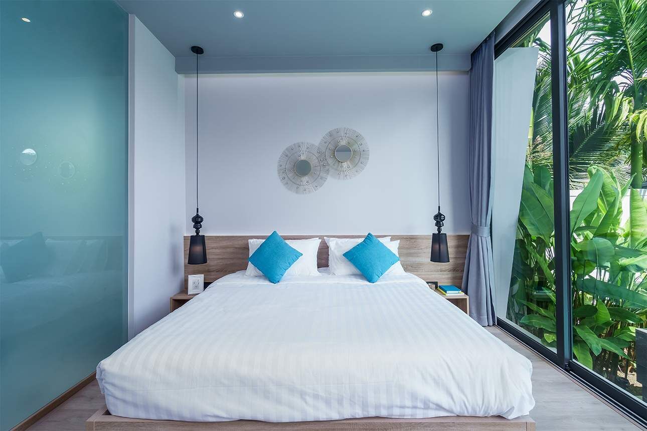 Quarto moderno com uma cama grande, duas almofadas azuis, cabeceira de madeira e vista para o verde através de uma janela ampla.