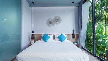 Quarto moderno com uma cama grande, duas almofadas azuis, cabeceira de madeira e vista para o verde através de uma janela ampla.