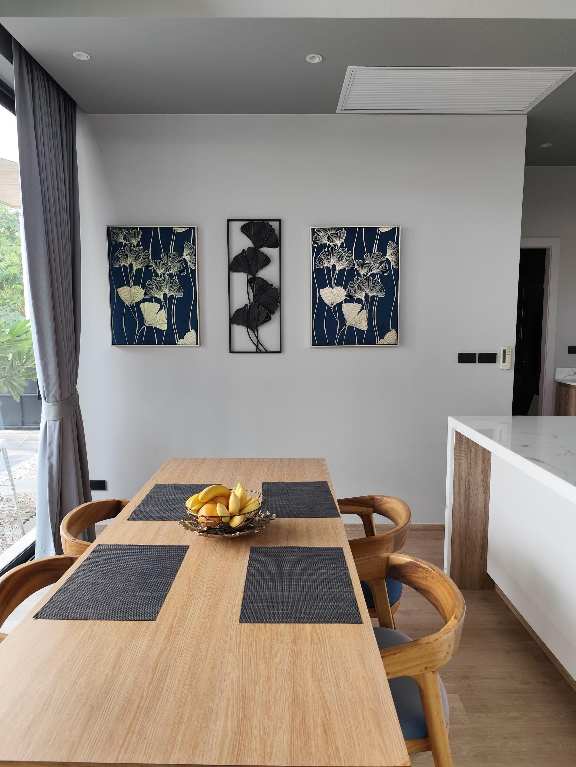 Uma área de refeições moderna com uma mesa de madeira, jogos americanos pretos e uma cesta de frutas. Três obras de arte fixadas na parede retratam designs florais abstratos.