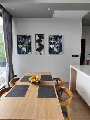 A modern dining area with a wooden table, black placemats, and a fruit bowl. Three wall-mounted artworks depict abstract floral designs. - Stylish 2BR Naiton Aileen Villa w/ Private Pool (Sa Khu)