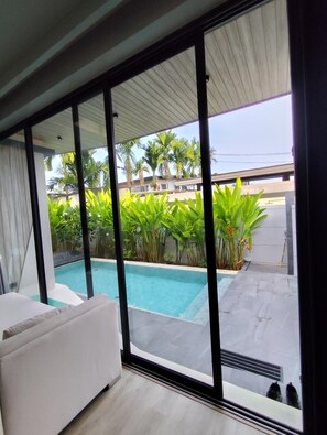 A modern room with a large sliding glass door leading to a swimming pool area, surrounded by lush greenery and palm trees. - Stylish 2BR Naiton Aileen Villa w/ Private Pool (Sa Khu)
