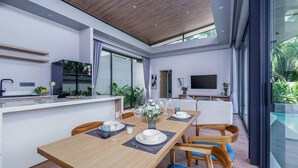 Een moderne eetruimte met een houten tafel voor vier personen, een keuken met een televisie, en een groot raam met uitzicht op een zwembad.