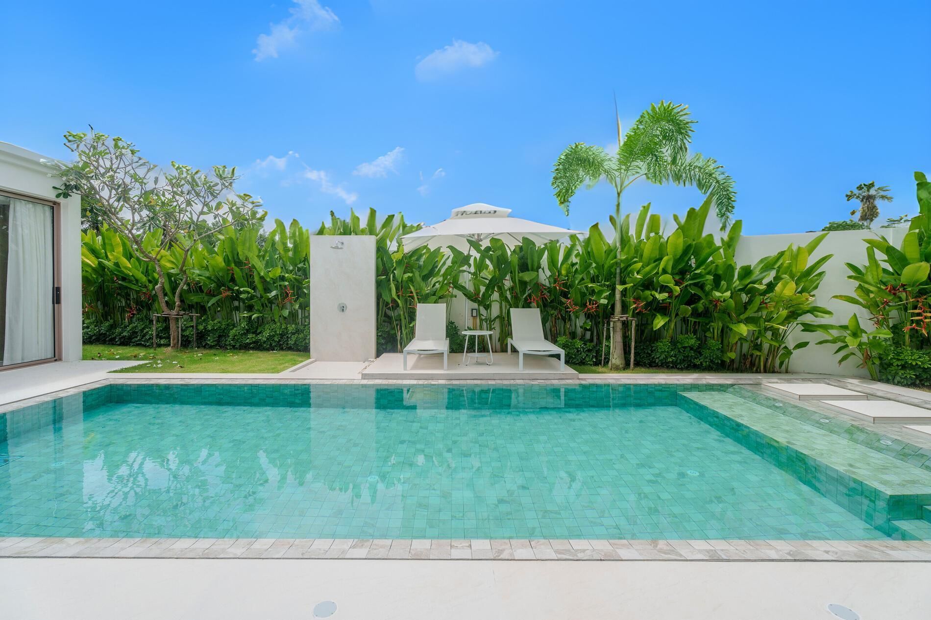 A rectangular swimming pool with clear blue water, surrounded by lush greenery and a white umbrella structure.