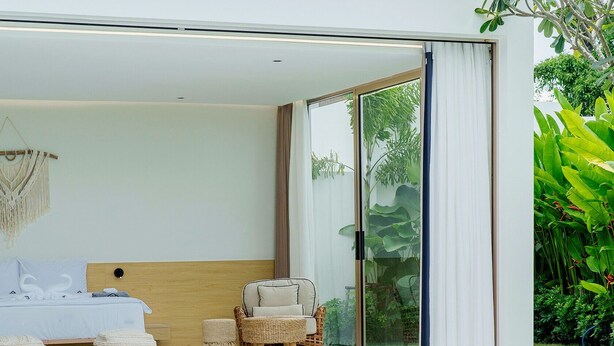 Een moderne slaapkamer met een groot bed, een stoel en een klein tafeltje. Er is een glazen schuifdeur naar een balkon met een zwembad.