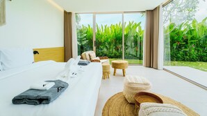 Een moderne hotelkamer met een groot bed, een klein zithoekje en uitzicht op een weelderige tuin door middel van ramen van de vloer tot aan het plafond.