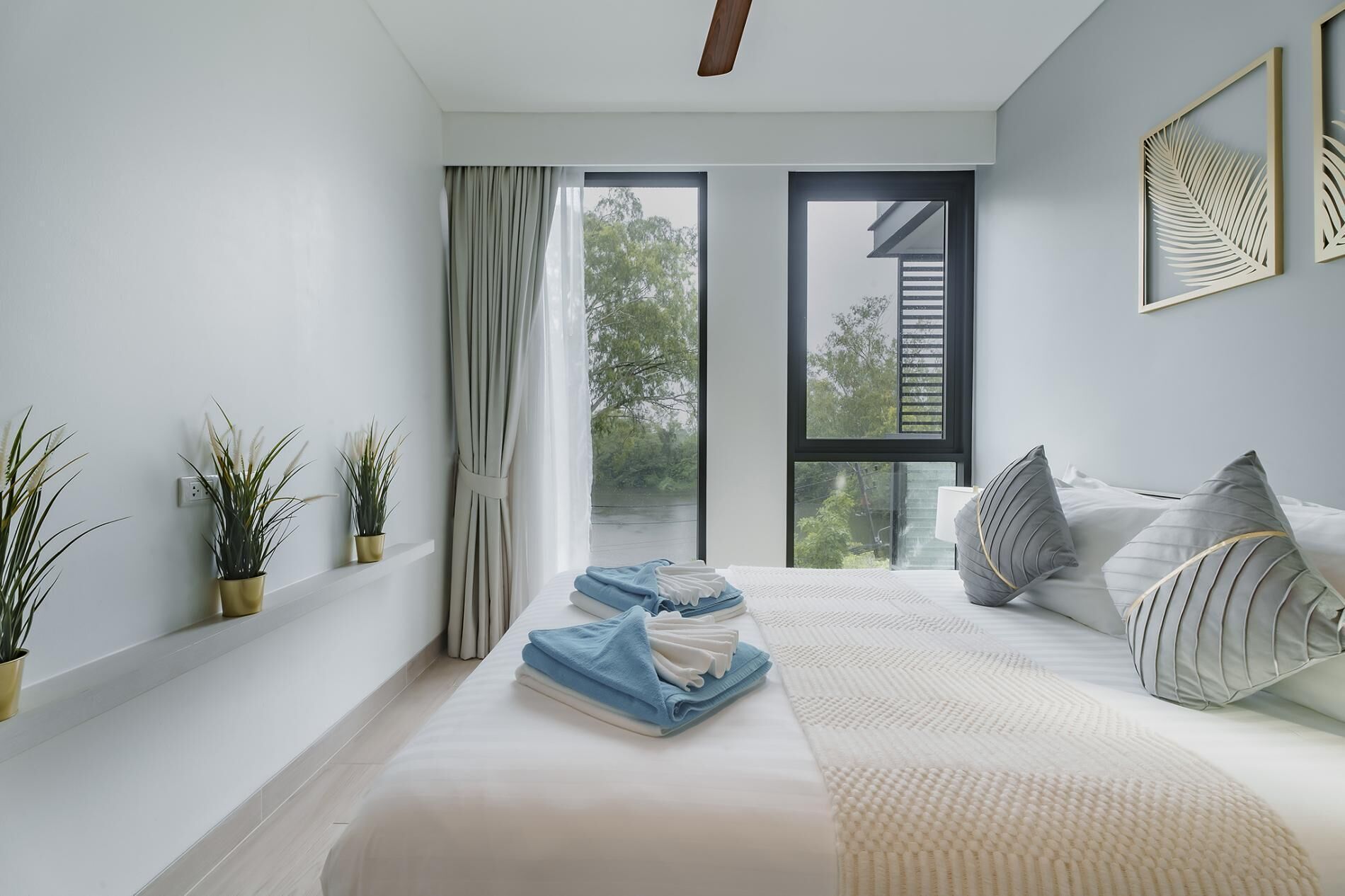 A modern bedroom with a large window, a bed with pillows, and two potted plants on a shelf.