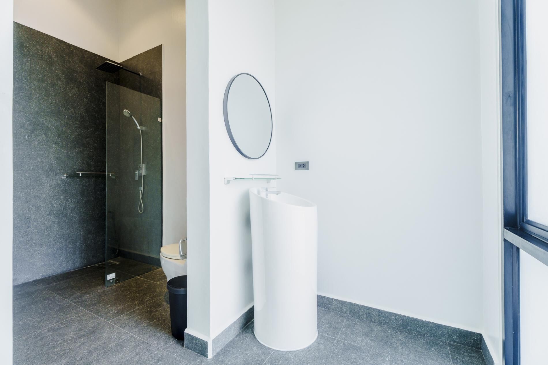 A modern bathroom with a glass shower enclosure, a white pedestal sink, and a round mirror.