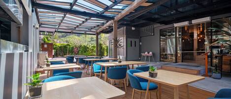 An indoor-outdoor dining area with wooden tables and blue chairs, a glass ceiling, and a view of greenery.