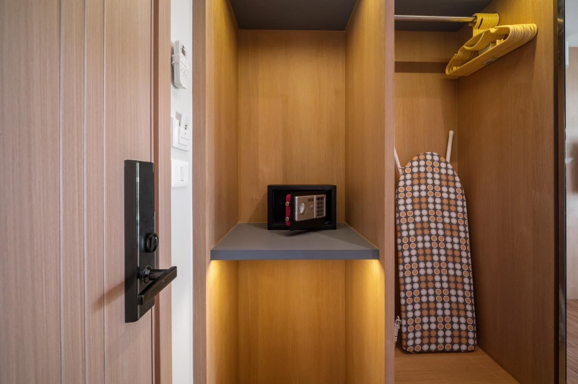 A compact room with a wooden wardrobe, a small safe, and a patterned ironing board.