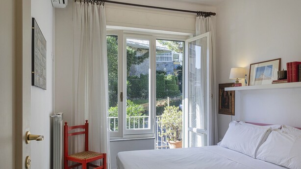 A bedroom with a bed, a chair, a window with curtains, a radiator, and a wall-mounted shelf with books and a picture. - S Margherita Harbour Apartment by Wonderful Italy (Santa Margherita Ligure)