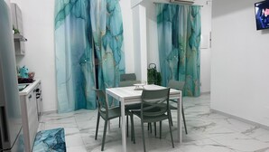 Ein Essbereich mit Tisch und Stühlen, eine Küche mit Kühlschrank und blaustichiges Vorhanggewebe.