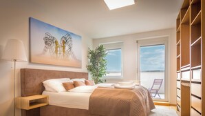 Ein Schlafzimmer mit Bett, Nachttisch, Lampe, Kunstwerk, Fenstern und Blick ins Freie.